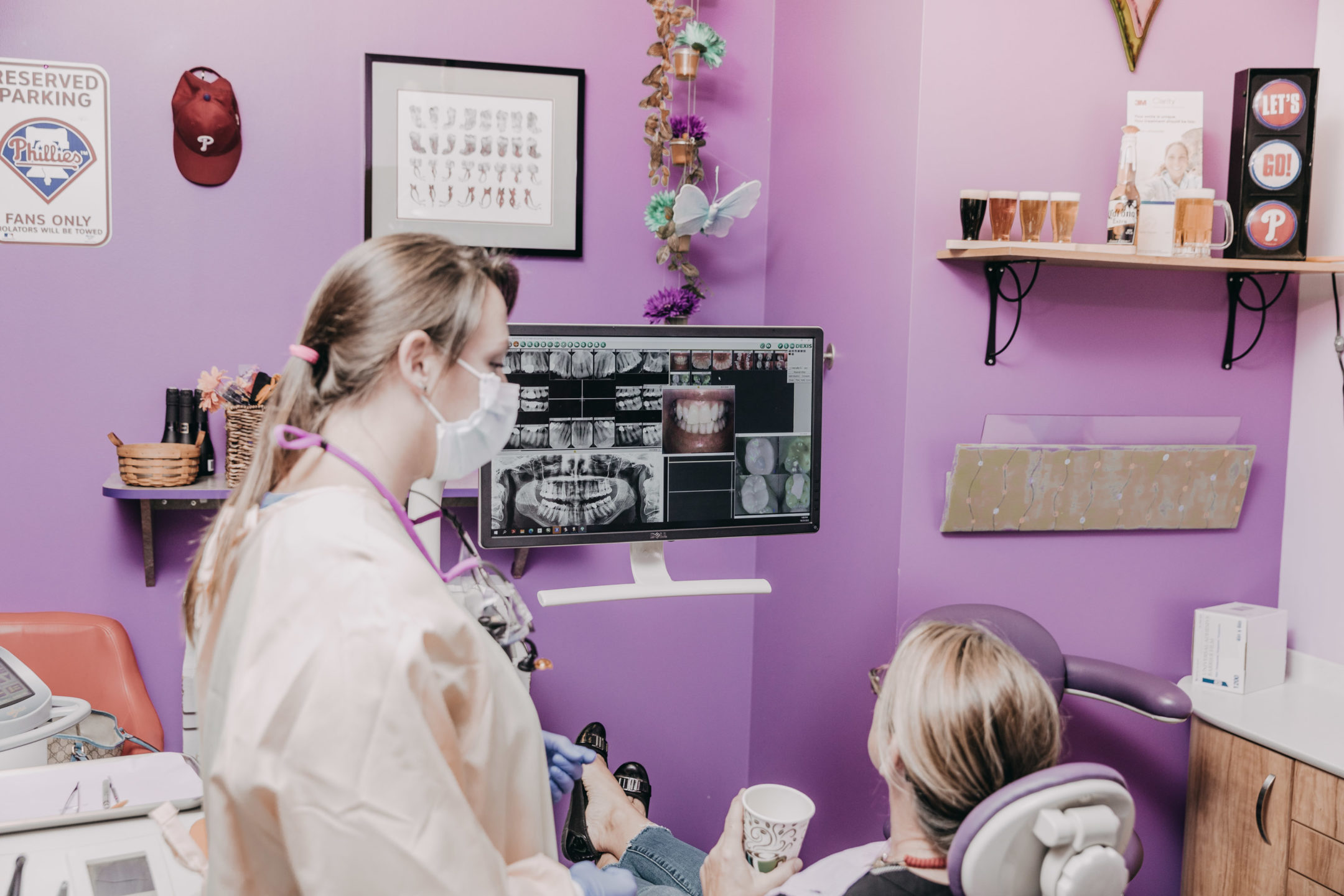  A dentist shows a patient her teeth on a screen as she sits in the dental chair with a cup of coffee. 