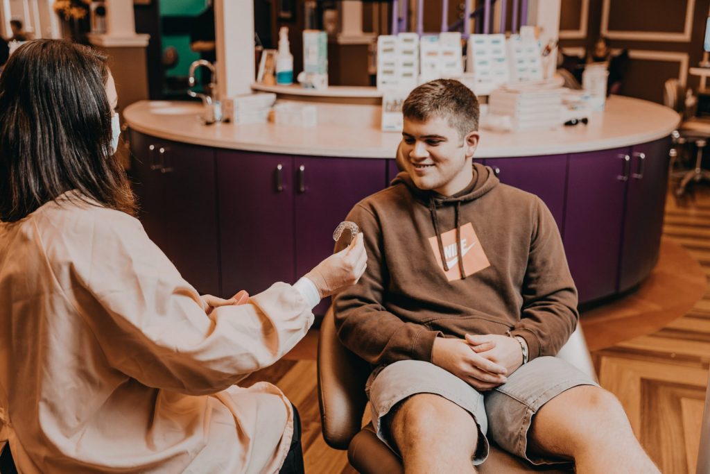 A female orthodontist speaks with a male patient about clear braces for adults, holding up a sample product. 