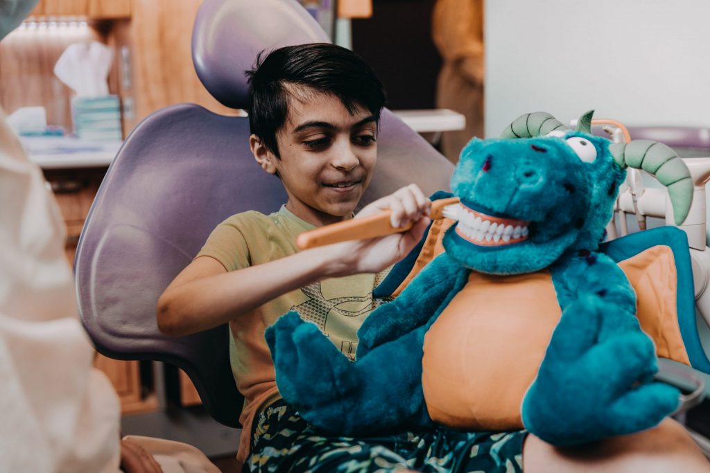 A young male dental patient brushes a dragon puppet’s teeth with an oversized toothbrush. 