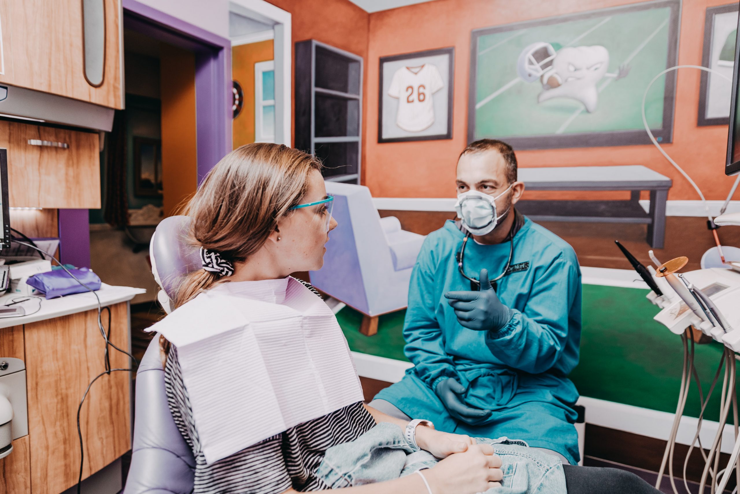 Male dentist wearing facemask and personal protective equipment (PPE) talks to female patient during dentist appointment.