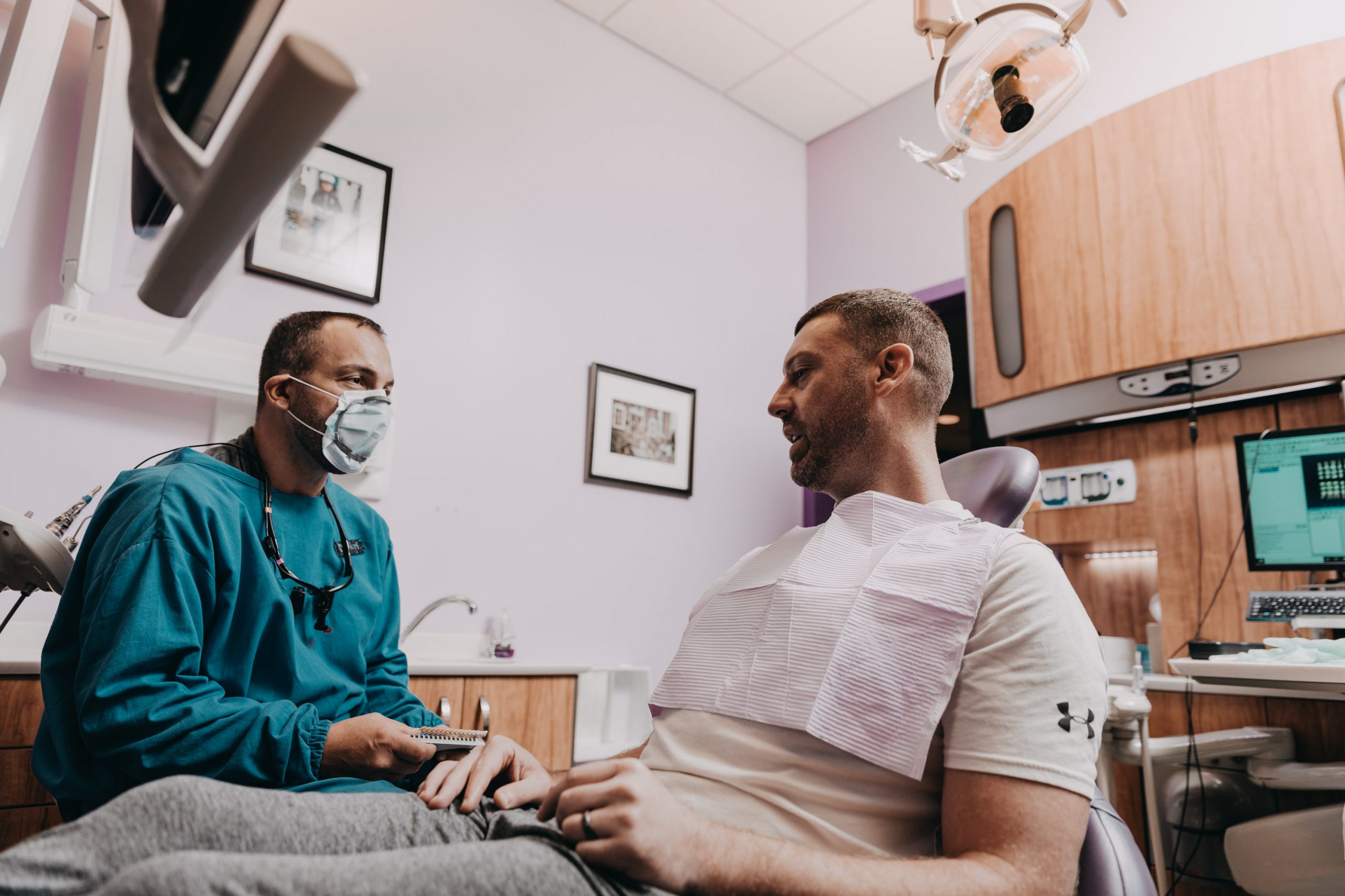  Male dentist wearing facemask and personal protective equipment (PPE) talks to male patient during dentist appointment.