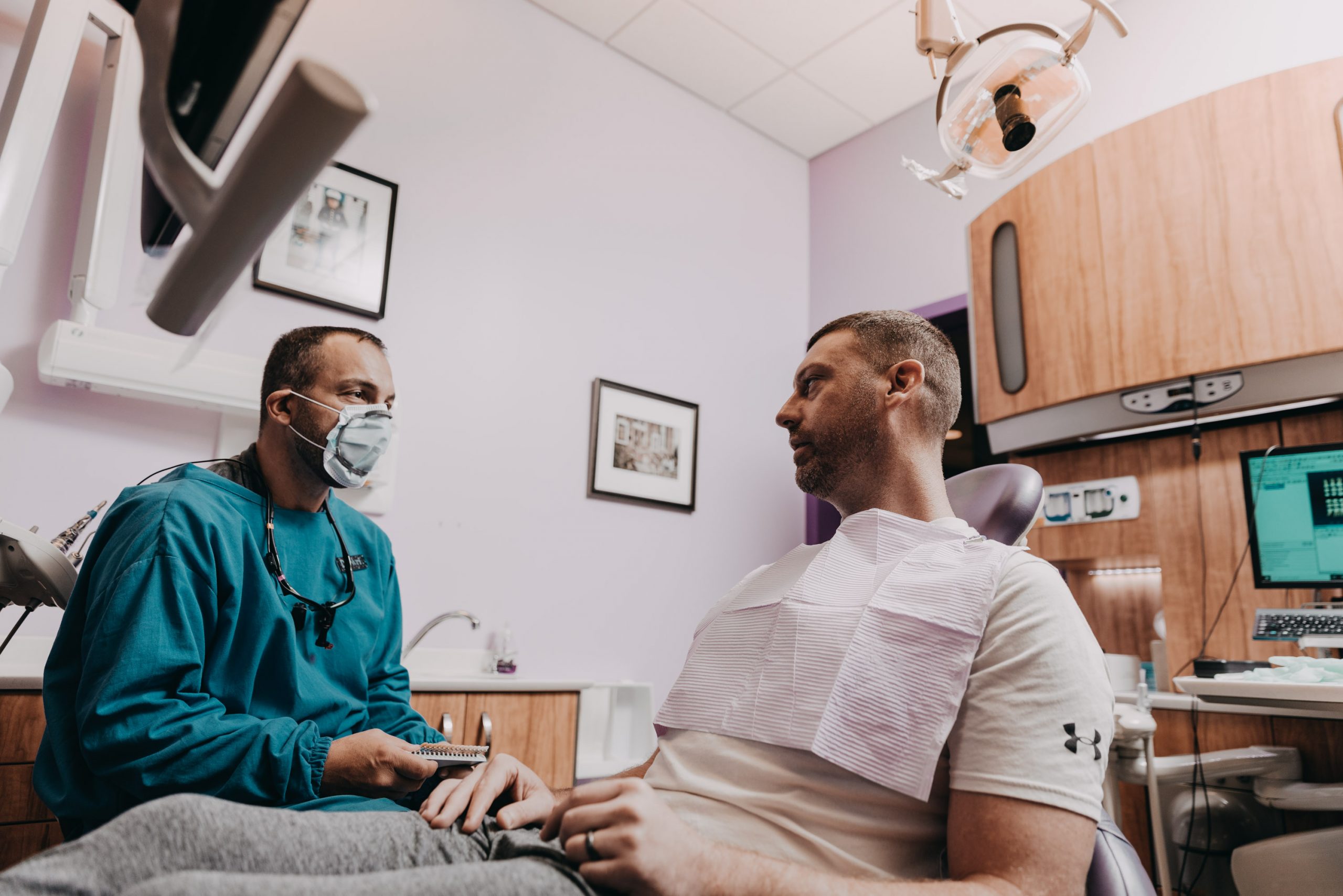 A male adult dentist in green safety gown and facemask discusses dental treatment with a male patient. 