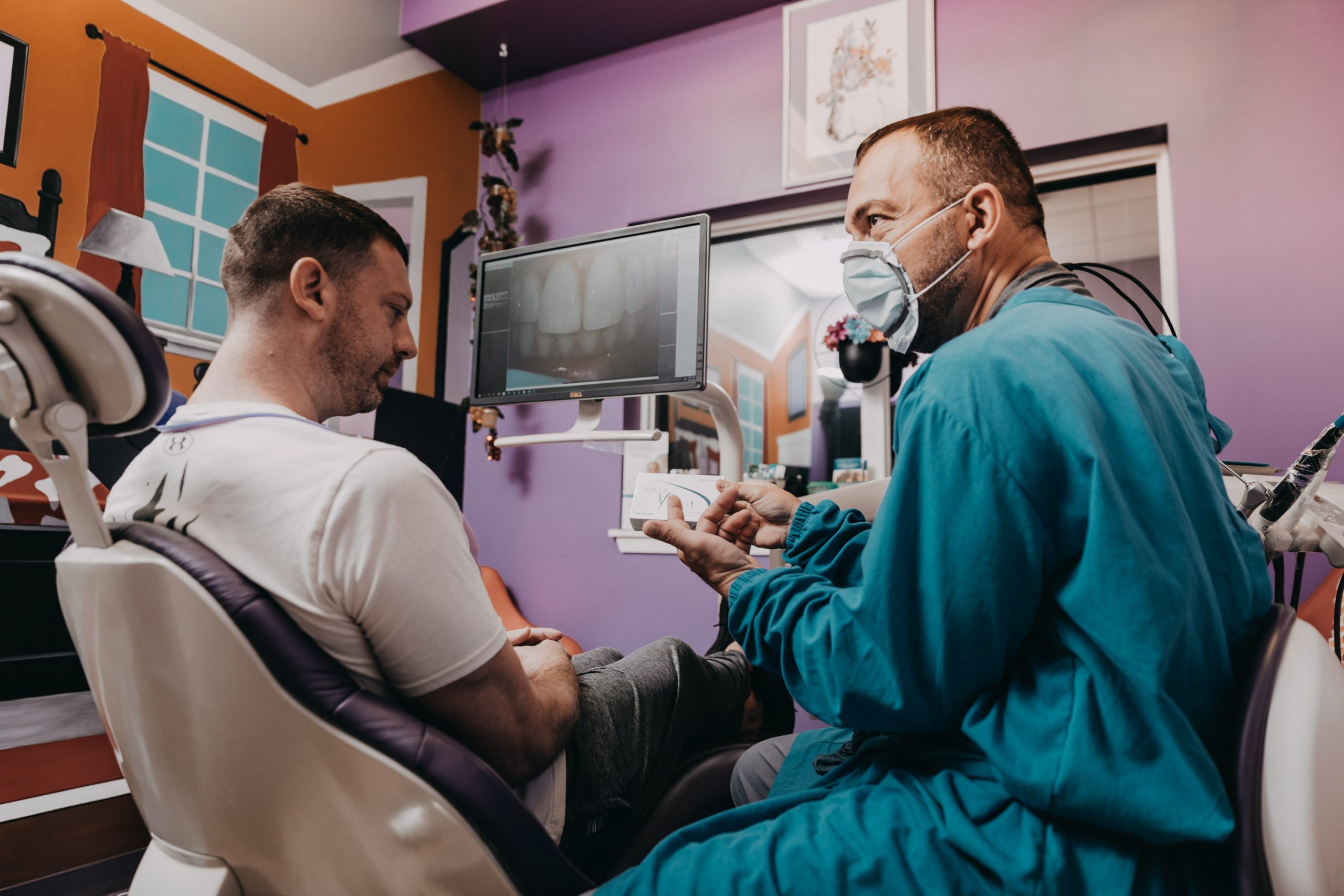 A male dentist wearing personal protective equipment (PPE) explains dental work options to a male patient.. 