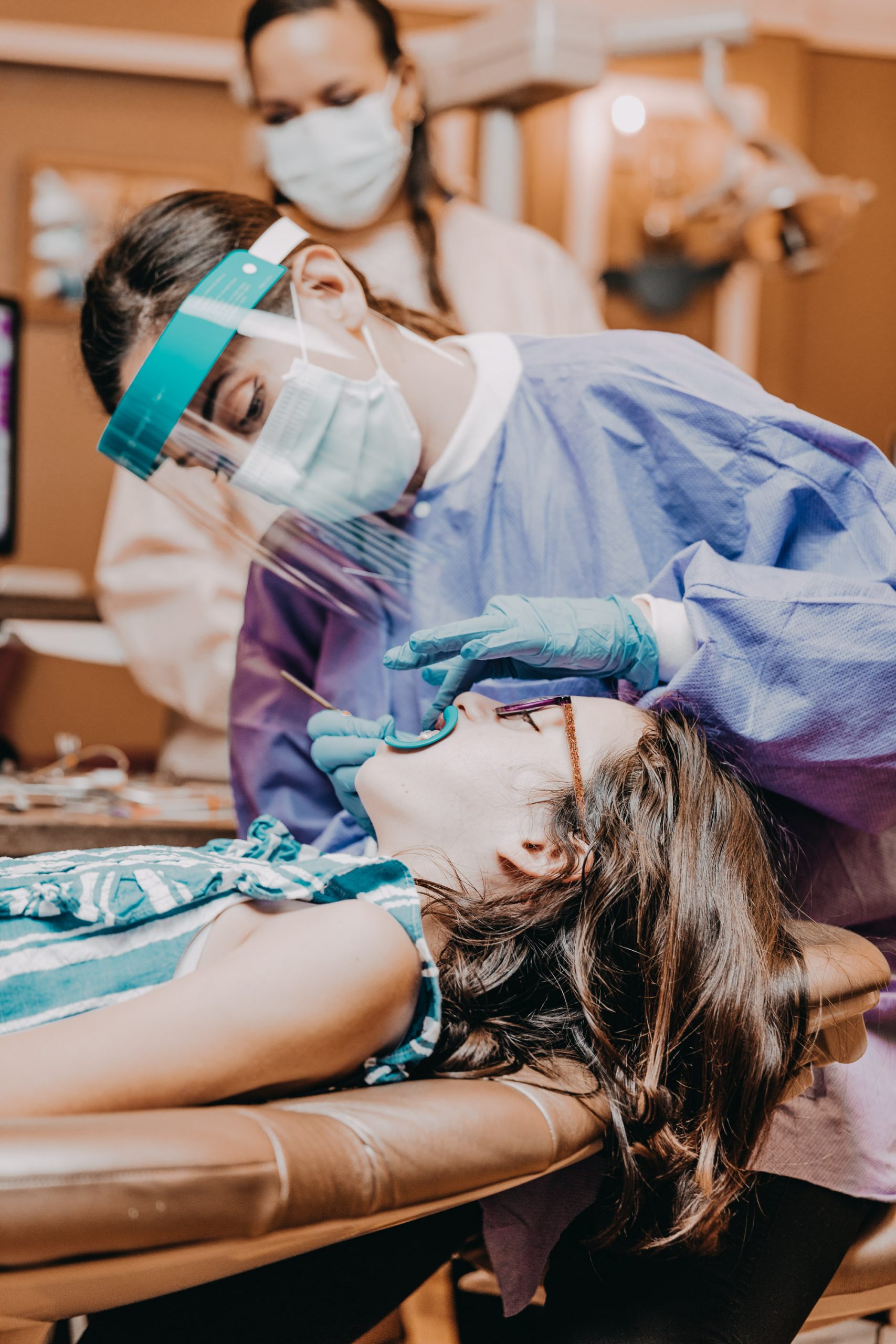 Female orthodontist wearing full, blue personal protective equipment (PPE) performs orthodontic work on young female. 