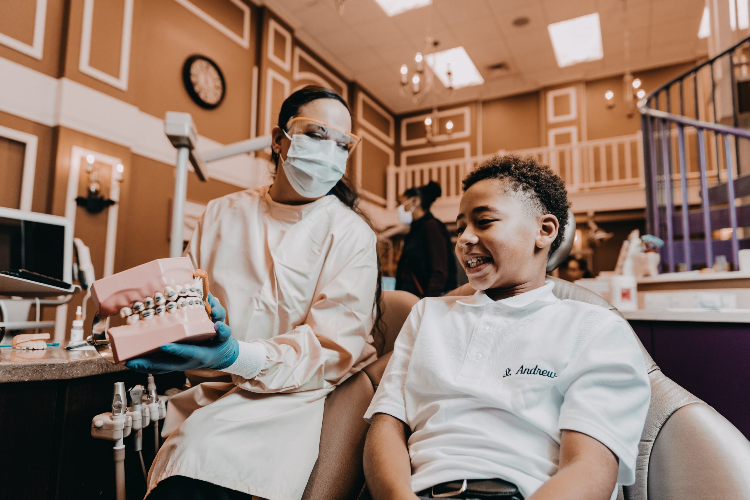  Female pediatric dental specialist shows young black male dental patient a large model of a mouth. 