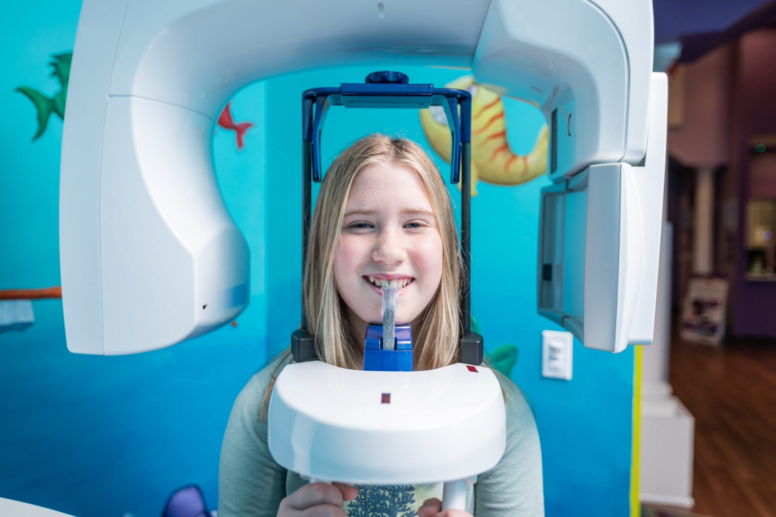  Young female child with blonde hair stands in a digital dental X-ray machine with sea-themed background.