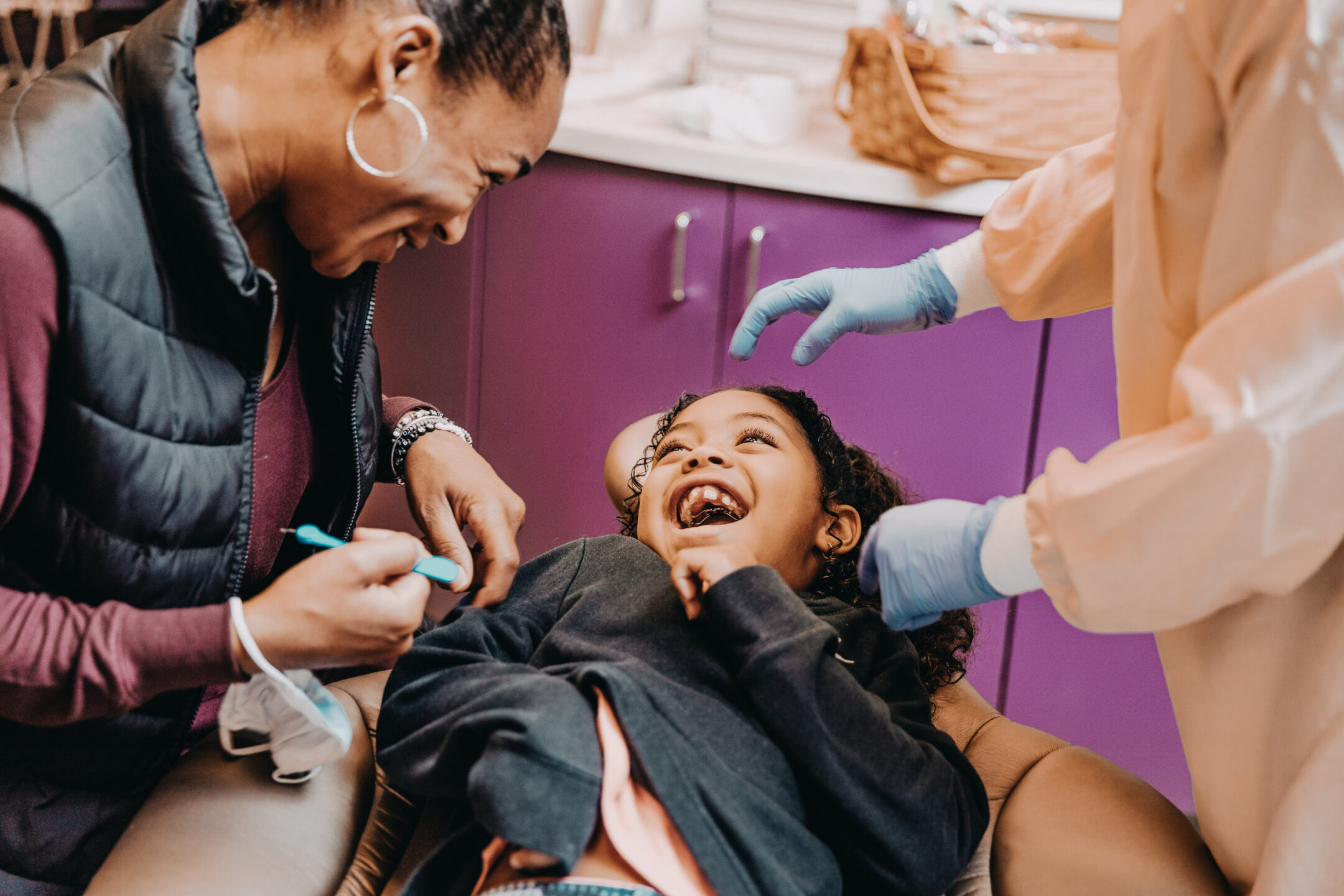 A girl lies back in a dental exam chair and giggles while a dentist and assistant lean over her smiling. 