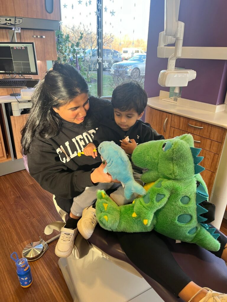 A baby sits on his mom’s lap in a dental chair with a stuffed dragon and shark toy.
