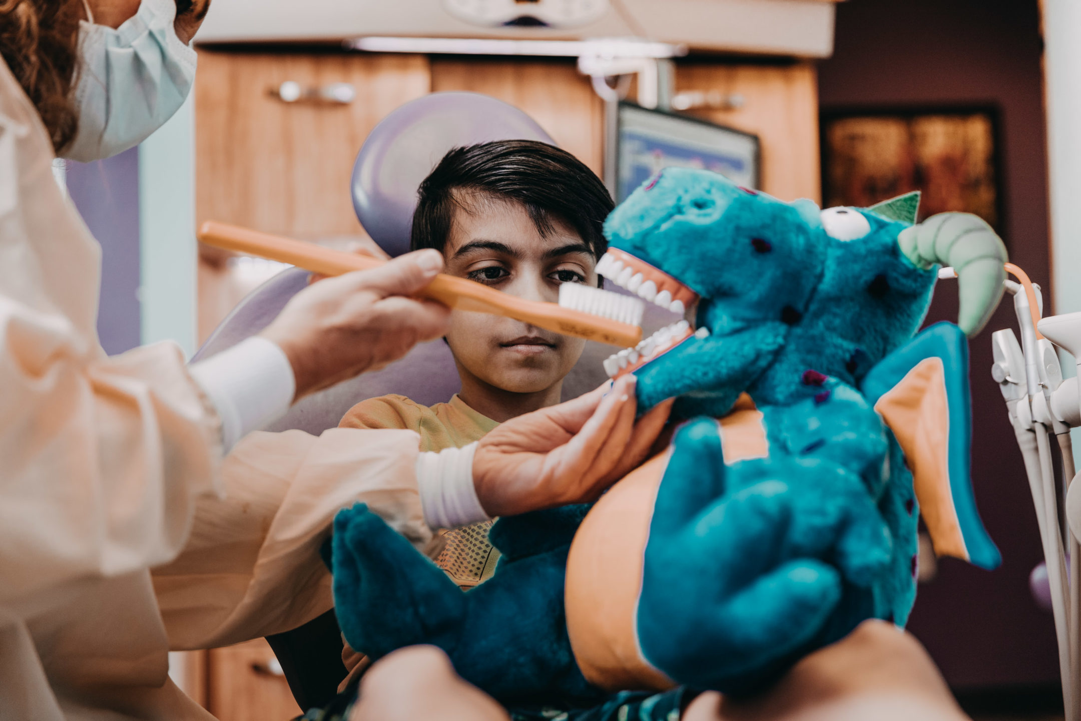 Dentist shows a young boy how to properly brush teeth using a plush mascot for demonstration