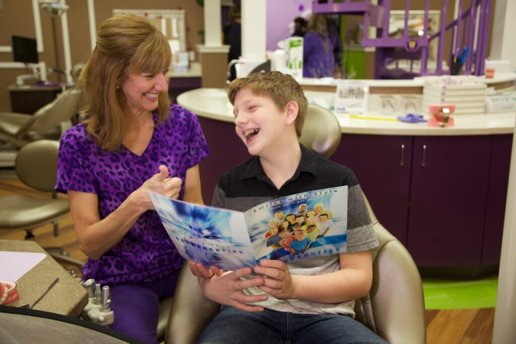Image of dental assistant in purple uniform laughing with young male patient holding a pamphlet on orthodontics.