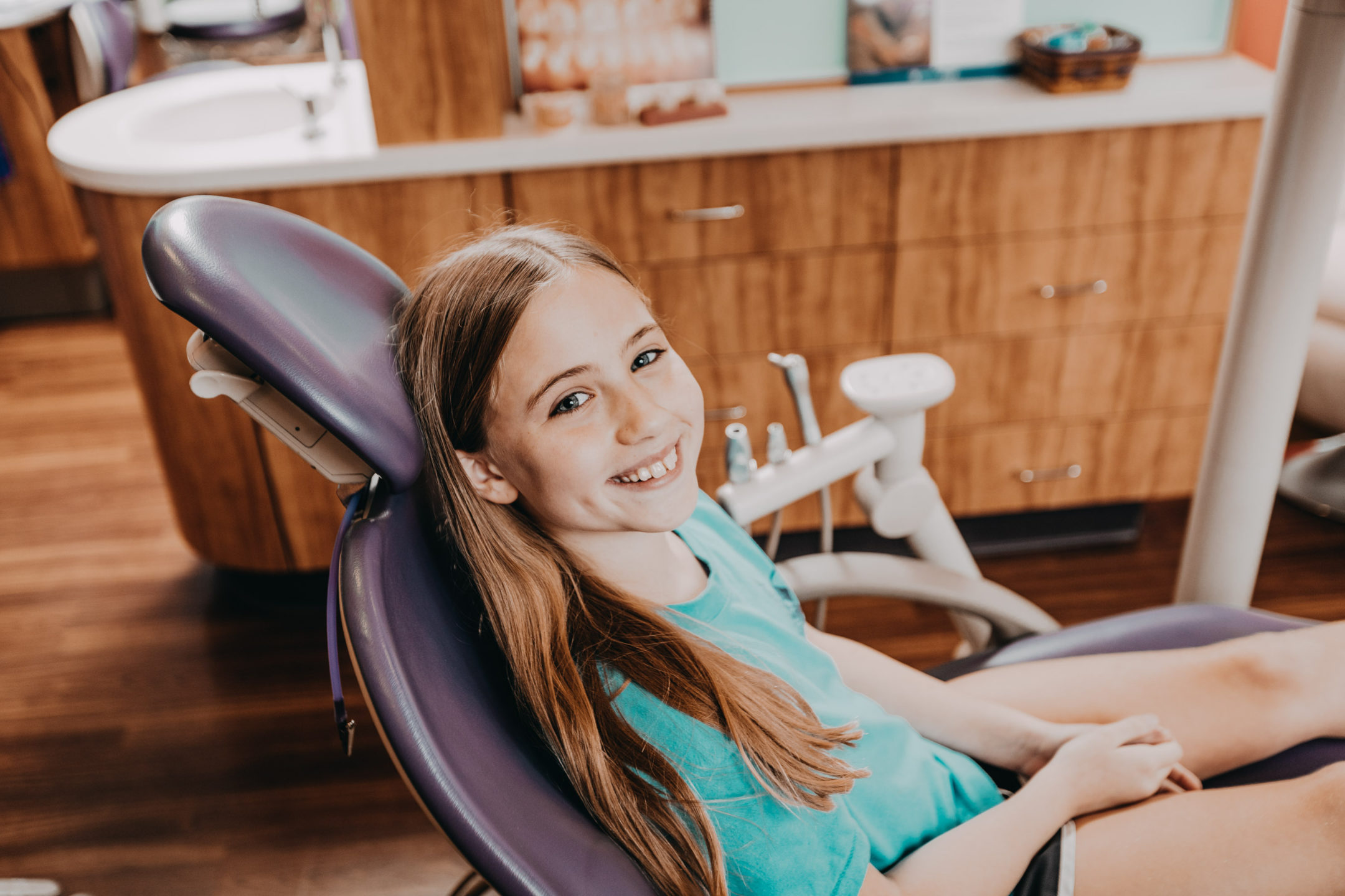 Smiling young girl sits in a dentist chair and looks at camera in a wram, wooden dentist office