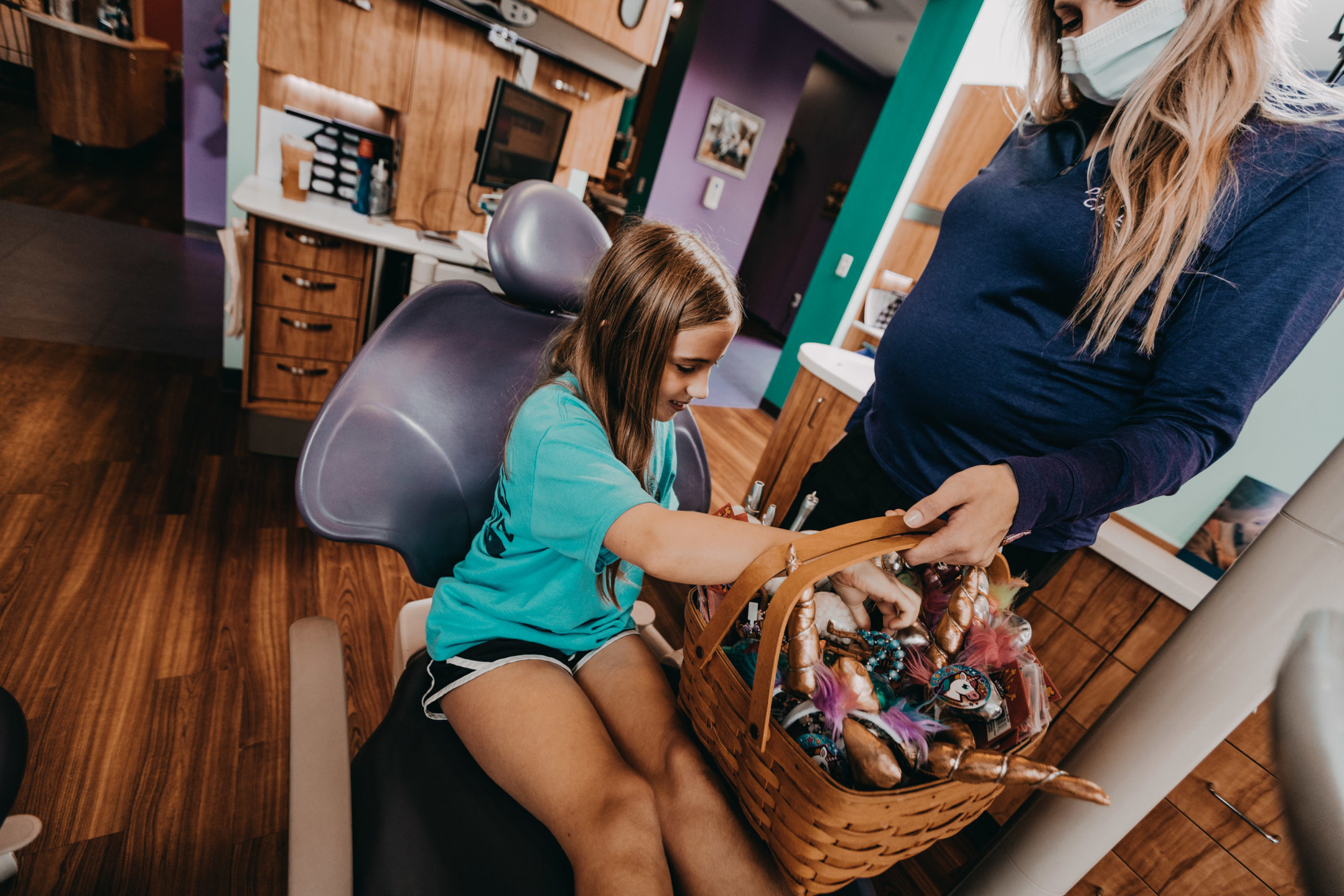 A woman holds a basket of toys while a young dental patient picks one. 