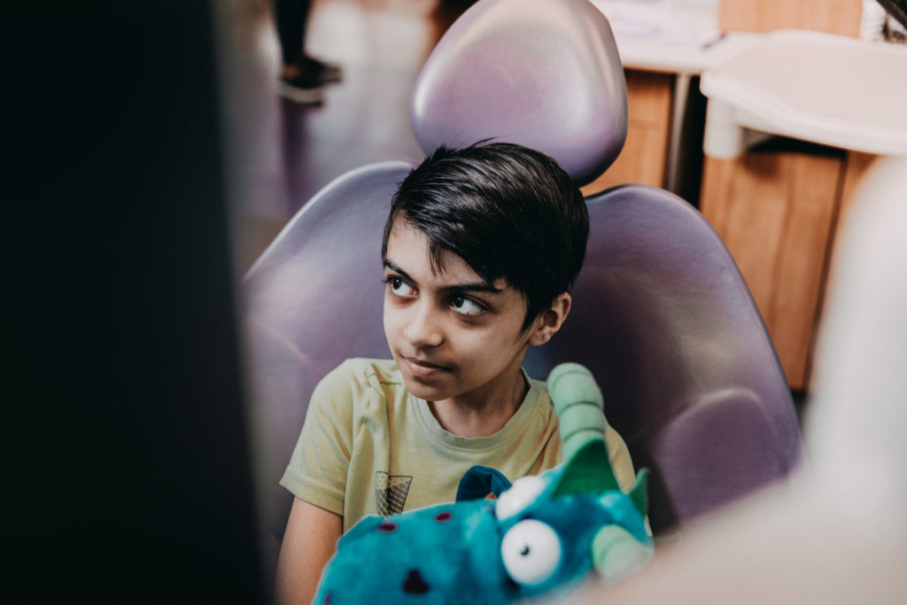 A young boy sits in a dental exam chair with a stuffed dragon toy. 