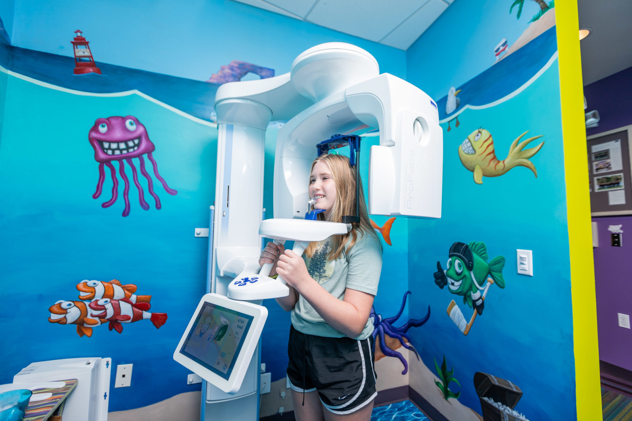  A girl stands in a dental X-ray machine in a brightly-decorated room. 