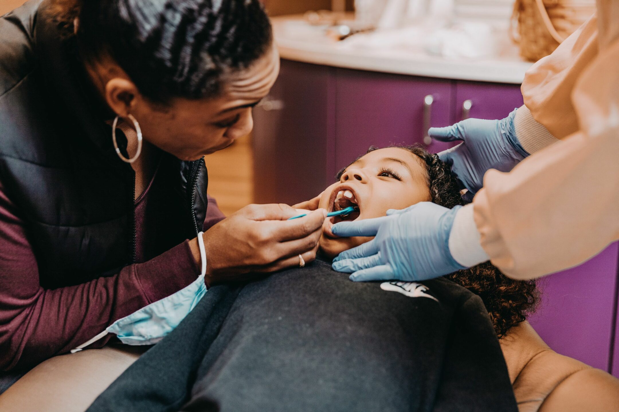  A dentist helps a mother look at her daughter’s teeth while the girl sits in a dental chair. 