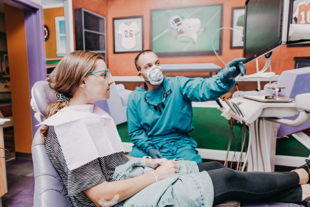 A dentist sits next to a patient in an exam chair and points at a screen.