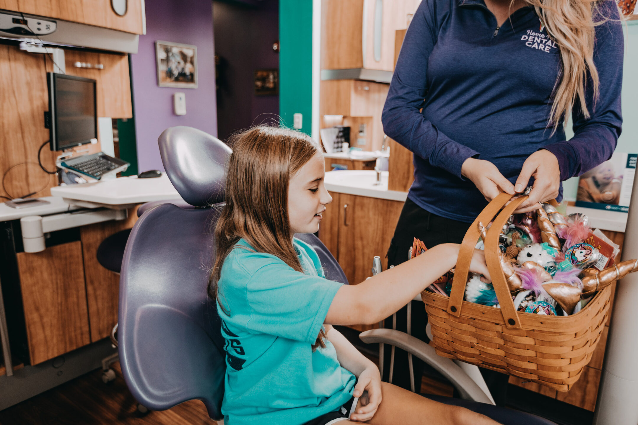 A girl sits in a dental chair as she picks out a toy from a basket held by a staff member. 