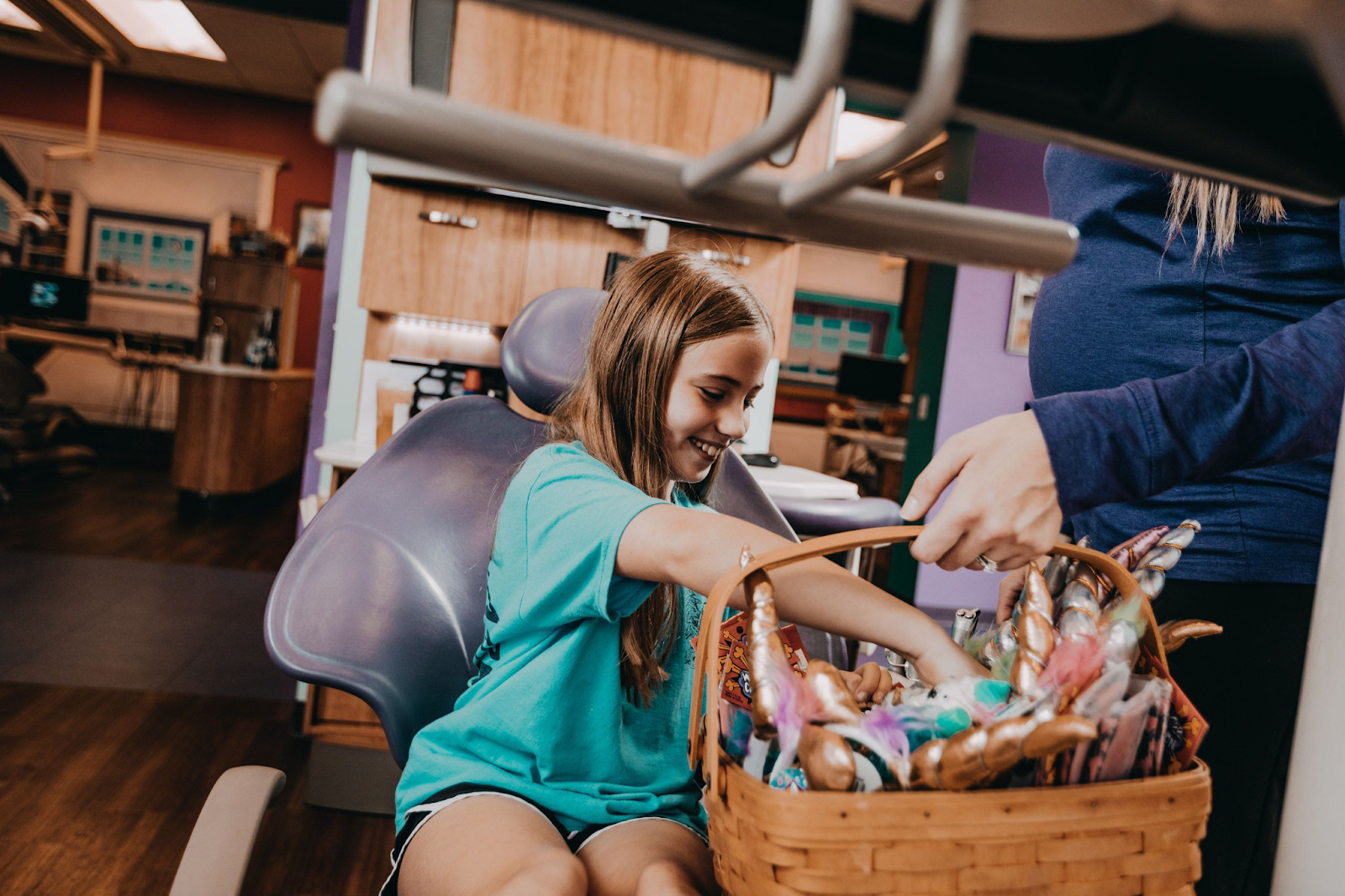 A girl sits in a dental exam chair and picks a prize from a basket. 