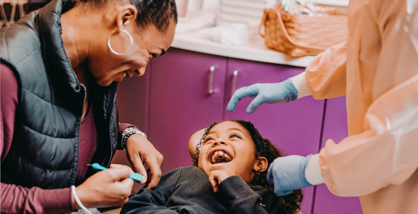 A girl laughs in a dental exam chair as her mom and a dentist stand next to her. 