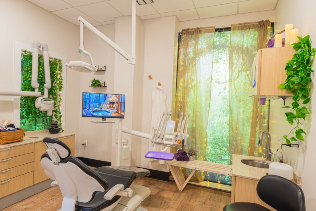  Image of a room in an adult dental practice with a forest theme, large-screen TV, and leaf-covered window.