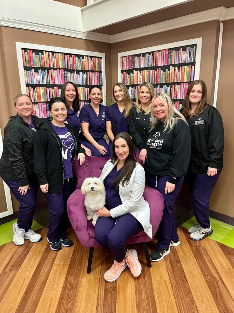 Photo of the Newtown Dentistry orthodontic team with bookshelves behind them and a white dog sitting on a purple chair. 