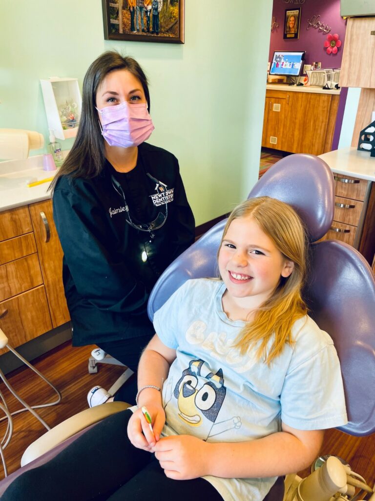 Photo of young smiling girl sitting in a purple dental chair with a dentist sitting beside her.