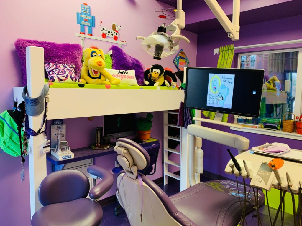 Image of a kids’ dental space with purple chairs and walls, a large-screen TV, and stuffed animals on a shelf. 